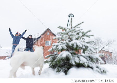 Two men jump up in winter in a snowdrift near the Christmas tree and a dog spoils the frame. Front view. 95013180