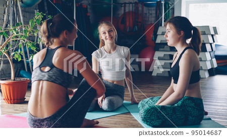 Young female yoga students are sitting on mats and talking while waiting for morning practice in large light gym. Fitness balls, step aerobic platforms and other sports equipment is visible. Young female yoga students are sitting on mats and talking while waiting for morning practice in large light gym. Fitness balls, step aerobic platforms and other sports equipment is visible. 95014687