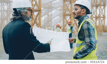 Back view of engineers discussing construction project looking at blueprint while surveyors are busy with equipment in background. Building and management concept. 95014724