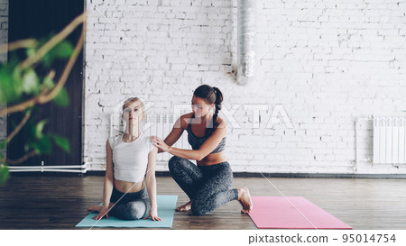 Cheerful attractive yoga teacher is helping new student master cow face pose during one-to-one class in modern sports center. Women are smiling and laughing, friendly atmosphere. 95014754