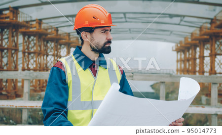 Portrait of serious foreman wearing uniform checking blueprint and looking at construction site outdoors in urban area. People and occupation concept. Portrait of serious foreman wearing uniform checking blueprint and looking at construction site outdoors in urban area. People and occupation concept. 95014773