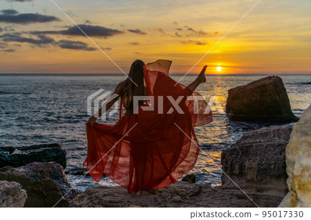 a dancing woman in a red flying dress on the ocean or on the sea beach against the backdrop of the sunset sky. 95017330