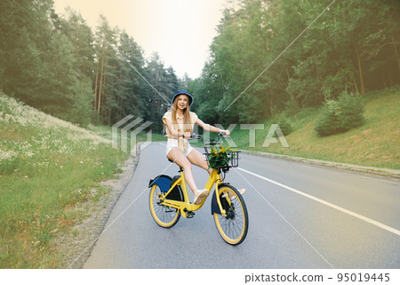 An attractive young tourist rides a bicycle on a road near the forest, stretching her legs and having fun and excited expressions, in the open air. An attractive young tourist rides a bicycle on a road near the forest, stretching her legs and having fun and excited expressions, in the open air. 95019445
