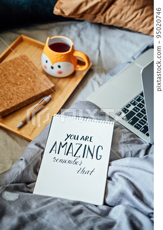 Positive daily affirmations for self love. Words You Are Amazing in notebook near laptop and tea coffee cup at bad in home. Handwritten affirmations text You Are Amazing in notepad 95020746