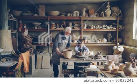 Experienced potter loving prandparent is teaching his young grandchild pottery in small home workshop. Boy is working with throwing wheel, grandpa is talking to him. 95022241