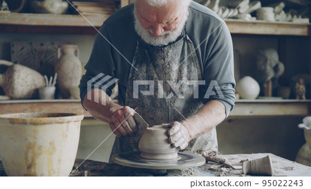 Senior male potter is working at bottom part of ceramic pot on spinning potter's wheel in workplace. Creating traditional eathenware and conventional pottery concept. 95022243