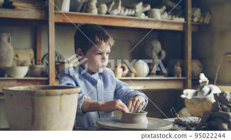 Cute curious child is making clay pot on spinning throwing wheel in grandfather's professional workshop. Brown ceramic vases, handmade pots, potter's tools are visible. Cute curious child is making clay pot on spinning throwing wheel in grandfather's professional workshop. Brown ceramic vases, handmade pots, potter's tools are visible. 95022244