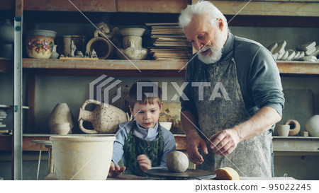 Caring silver-haired grandfather is teaching young cute grandson to work with clay on throwing-wheel in small cozy workshop. Pottery, family hobby and handicraft concept. Caring silver-haired grandfather is teaching young cute grandson to work with clay on throwing-wheel in small cozy workshop. Pottery, family hobby and handicraft concept. 95022245