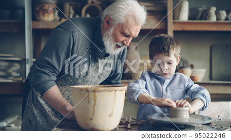 Concentrated young boy is molding clay into ceramic pot on spinning throwing wheel and his loving experienced grandfather is talking to him. Pottery and family tradition concept. Concentrated young boy is molding clay into ceramic pot on spinning throwing wheel and his loving experienced grandfather is talking to him. Pottery and family tradition concept. 95022246