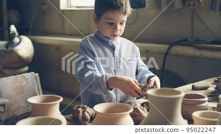 Curious little boy is playing with clay at working table in potter's workshop, kneading clay and tearing into pieces. Happy childhood, hobby, new experience concept. Curious little boy is playing with clay at working table in potter's workshop, kneading clay and tearing into pieces. Happy childhood, hobby, new experience concept. 95022247