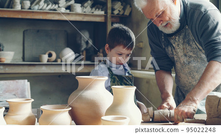 Experienced male potter is mixing and kneading clay while his cute young grandson is helping him in little workshop. Pottery, family tradition and childhood concept. Experienced male potter is mixing and kneading clay while his cute young grandson is helping him in little workshop. Pottery, family tradition and childhood concept. 95022248