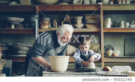 Curious little boy is learning pottery from his experienced grandfather in small home studio. Child is forming clay to make pot on potter's wheel, his granddad is helping him. 95022249