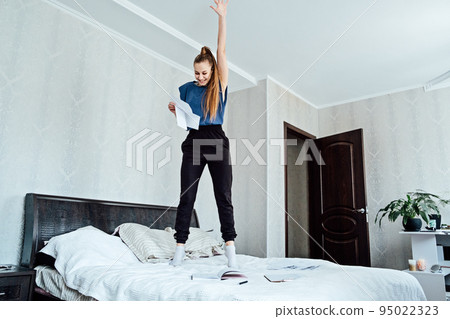 Excited cheerful young woman received a letter, she is hired, jumping on sofa at home, getting good news, feeling joy, dancing, laughing, making winner gesture, happy to win prize Excited cheerful young woman received a letter, she is hired, jumping on sofa at home, getting good news, feeling joy, dancing, laughing, making winner gesture, happy to win prize 95022323