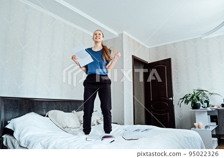 Excited cheerful young woman received a letter, she is hired, jumping on sofa at home, getting good news, feeling joy, dancing, laughing, making winner gesture, happy to win prize Excited cheerful young woman received a letter, she is hired, jumping on sofa at home, getting good news, feeling joy, dancing, laughing, making winner gesture, happy to win prize 95022326