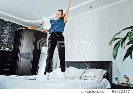 Excited cheerful young woman received a letter, she is hired, jumping on sofa at home, getting good news, feeling joy, dancing, laughing, making winner gesture, happy to win prize Excited cheerful young woman received a letter, she is hired, jumping on sofa at home, getting good news, feeling joy, dancing, laughing, making winner gesture, happy to win prize 95022327
