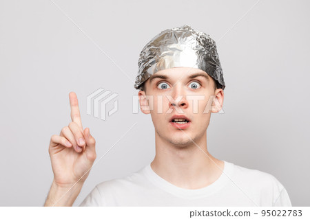 Paranoid young man wearing tin foil hat pointing up studio shot on gray background 95022783