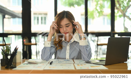 Frustrated female worker holding her head and looking at financial documents, thinking find solution problem of work 95022961