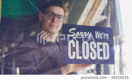 Handsome male cafe worker in apron is changing doorplate to sorry we are closed. End of work day, evening and catering business concept. 95026016