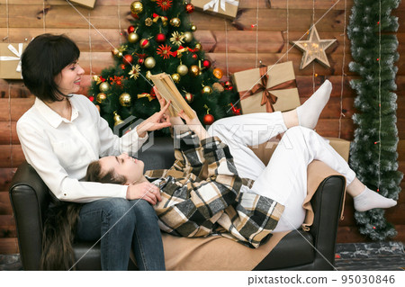 Mom and daughter are sitting comfortably on the couch reading fairy tales on New Year's Eve Mom and daughter are sitting comfortably on the couch reading fairy tales on New Year's Eve 95030846