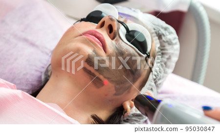 A woman in protective glasses with a smeared black mask on her face is undergoing a procedure in a beauty salon A woman in protective glasses with a smeared black mask on her face is undergoing a procedure in a beauty salon 95030847