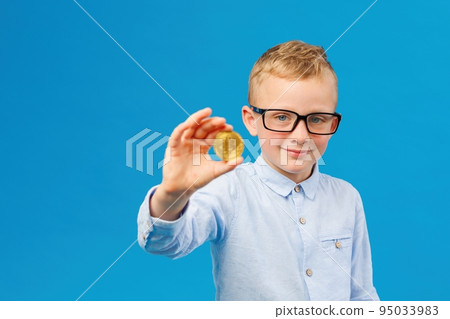 Cryptocurrency and modern finance. Cheerful boy in eyeglasses holding golden bitcoin in studio on blue background. 95033983