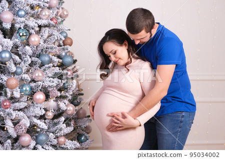 Husband and wife enjoy the pregnancy together, standing near the Christmas tree. Happy baby wait and Christmas Husband and wife enjoy the pregnancy together, standing near the Christmas tree. Happy baby wait and Christmas 95034002
