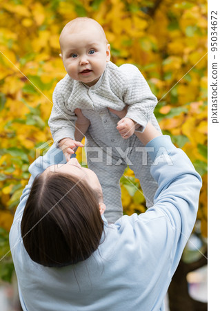 Woman lifts up charmed baby clenching fist in autumn park 95034672
