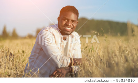 African American agriculturist sits on wheat field smiling 95034810