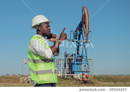 Black engineer talks on phone with colleague near jack-pump Black engineer talks on phone with colleague near jack-pump 95034855