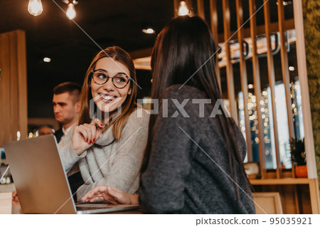 Two young business women sitting at table in cafe. Girl shows colleague information on laptop screen. Girl using smartphone, blogging. Teamwork, business meeting. 95035921