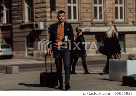 Going to airport terminal. Confident businessman traveler walking on city streets and pulling his suitcase drinking coffee and speaking on smartphone Going to airport terminal. Confident businessman traveler walking on city streets and pulling his suitcase drinking coffee and speaking on smartphone 95035925