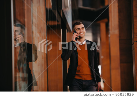 Going to airport terminal. Confident businessman traveler walking on city streets and pulling his suitcase drinking coffee and speaking on smartphone Going to airport terminal. Confident businessman traveler walking on city streets and pulling his suitcase drinking coffee and speaking on smartphone 95035929