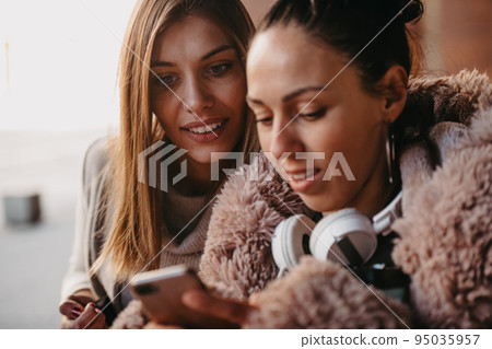Latino woman with her friend is smiling happily using a smartphone and headphones in the city. 95035957
