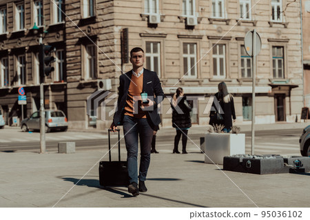 Going to airport terminal. Confident businessman traveler walking on city streets and pulling his suitcase drinking coffee and speaking on smartphone 95036102