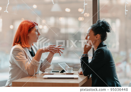 Two young business women sitting at table in cafe. Girl shows colleague information on laptop screen. Girl using smartphone, blogging. Teamwork, business meeting. 95036448