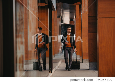 Going to airport terminal. Confident businessman traveler walking on city streets and pulling his suitcase drinking coffee and speaking on smartphone Going to airport terminal. Confident businessman traveler walking on city streets and pulling his suitcase drinking coffee and speaking on smartphone 95036499