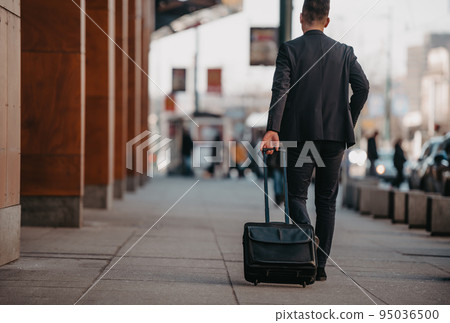 Going to airport terminal. Confident businessman traveler walking on city streets and pulling his suitcase drinking coffee and speaking on smartphone 95036500