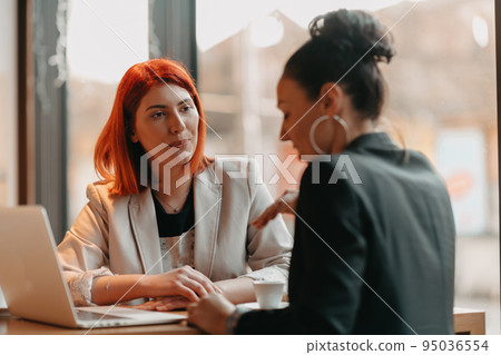 Two young business women sitting at table in cafe. Girl shows colleague information on laptop screen. Girl using smartphone, blogging. Teamwork, business meeting. Two young business women sitting at table in cafe. Girl shows colleague information on laptop screen. Girl using smartphone, blogging. Teamwork, business meeting. 95036554