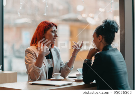 Two young business women sitting at table in cafe. Girl shows colleague information on laptop screen. Girl using smartphone, blogging. Teamwork, business meeting. 95036563