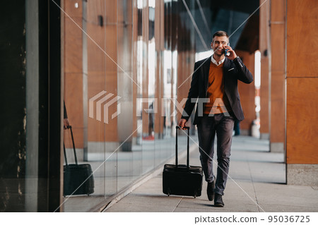 Going to airport terminal. Confident businessman traveler walking on city streets and pulling his suitcase drinking coffee and speaking on smartphone 95036725