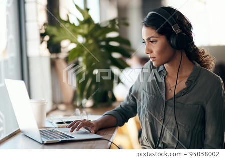 Female student working on laptop with headphones for a university project in a cafe, restaurant or coffee shop. Thinking and studying young university or college woman typing and listening to music 95038002