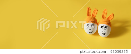 two white eggs in orange beanie with rabbit ears and cheerful faces on yellow background 95039255