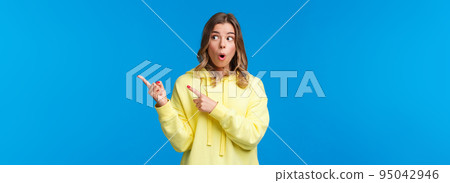Wow something interesting. Intrigued and surprised alluring blond woman in yellow hoodie talking about last rumors, pointing and looking right with impressed expression, blue background 95042946