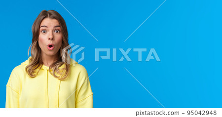 Close-up portrait of intrigued and curious good-looking young teenage girl with blond short hair, say wow, staring impressed and speechless at camera, standing blue background 95042948