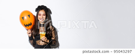 Image of cheerful asian girl trick or treating on halloween, holding sweets and orange balloon with scary face 95043297
