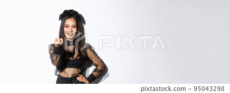 Beautiful woman in witch costume telling to come closer, flick finger and smiling, standing in halloween outfit, gothic makeup, standing over white background 95043298