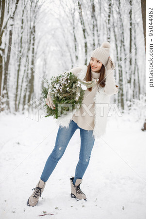 Young brunette woman standing in winter forest and holding a little Christmas tree. Female model posing for a photo. Woman wearing beige jacket and knitted hat. Young brunette woman standing in winter forest and holding a little Christmas tree. Female model posing for a photo. Woman wearing beige jacket and knitted hat. 95044962
