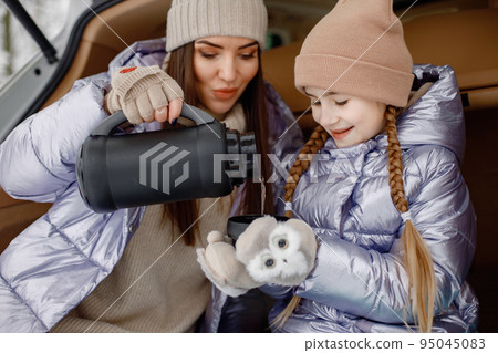 Young brunette woman sitting in open car's trunk with her daughter and holding a thermos with tea. Female models posing for a photo. Family wearing purple warm jackets and knitted hats. Young brunette woman sitting in open car's trunk with her daughter and holding a thermos with tea. Female models posing for a photo. Family wearing purple warm jackets and knitted hats. 95045083
