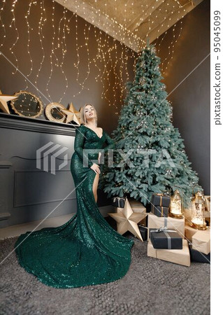 Portrait of woman posing for a photo. Gorgeous blonde girl in an elegant long green dress celebrates Christmas. Woman standing by a beautiful Christmas tree in expensive interiors. 95045099
