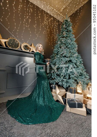 Portrait of woman posing for a photo. Gorgeous blonde girl in an elegant long green dress celebrates Christmas. Woman standing by a beautiful Christmas tree in expensive interiors. Portrait of woman posing for a photo. Gorgeous blonde girl in an elegant long green dress celebrates Christmas. Woman standing by a beautiful Christmas tree in expensive interiors. 95045102